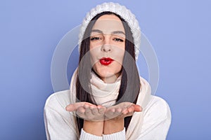 Young woman wearing winter clothes gesturing flying kiss against blue background, female with red pomade, girl dresses white cap