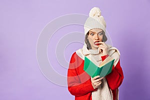 Young woman wearing winter hat isolated over purple background reading book