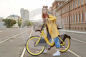 Young woman wearing yellow coat and colored pigtails, riding bike in city