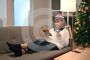 Young woman wears Christmas hat reading book and drinking red wine while lying on couch.