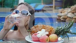 Young woman wears sunglassed drinks water while sunbathing in the pool at plate with fruits and plumeria flower in slow