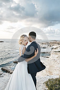 Young woman in a wedding dress and groom in a suit posing on the cliff near the ocean. Happy couple hugging together on
