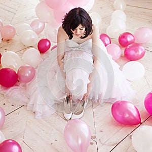 Young woman in wedding dress in luxury interior with a mass of pink and white balloons. Hold in hands her white shoes