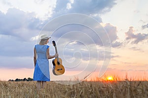 Young woman in wheat field with acoustic guitar at sunset