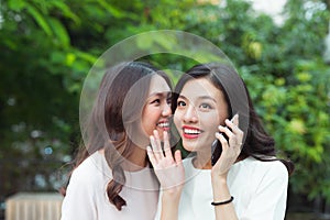 Young woman whispering into cheerful friend`s ear while on call