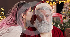 A young woman whispers something in the ear of a surprised Santa Claus sitting in a New Year's studio with lights, next