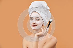 Young woman with white bath towel in her hair holding brush, applying face mask. Wellness and Spa concept on beige.