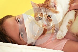 Young woman in white cloth virus face mask playing with her cat, detail on feline eyes, owner blurred in background