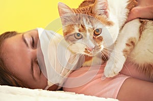 Young woman in white cloth virus face mask playing with her cat, detail on feline eyes, owner blurred in background