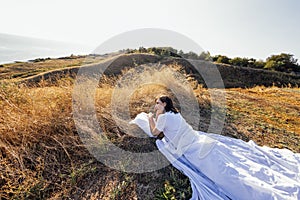 A young woman in white clothes lies on a white sheet and pillow in a field among dry grass. The girl relaxes and rests in nature photo