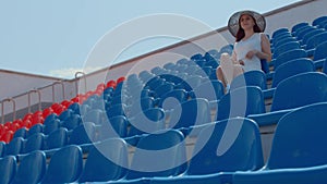 Young woman in in white dress and elegant hat looks away, sitting on stadium bleachers alone. Lonely spectator at street