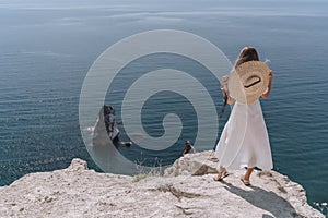A young woman in a white dress and hat stands with her back on a rock and looks at the sea. Rocks are visible in the sea. The