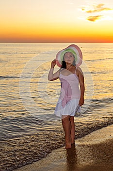 A young woman in a white dress and hat stands on the seashore at sunset photo