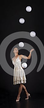 A young woman in a white dress juggles balls on a dark background