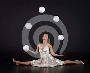 A young woman in a white dress juggles balls on a dark background