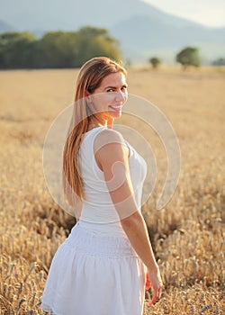 Young woman in white dress looking back over her shoulder, smiling, afternoon sun lit wheat field behind her