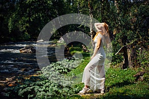 Young woman in white dress raised her face towards the sun. Dreamy pretty girl on the background of green forest and mountain