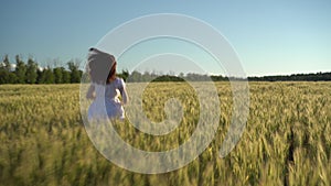 A young woman in a white dress runs along a green wheat field. The girl runs and raises her hands up. Back view.