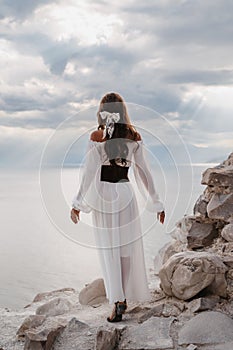 A young woman in a white dress stands barefoot on a cliff face in full height. The dress flutters in the wind. A sacred