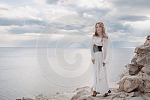 A young woman in a white dress stands barefoot on a cliff face in full height. The dress flutters in the wind. A sacred