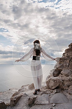 A young woman in a white dress stands barefoot on a cliff face in full height. The dress flutters in the wind. A sacred