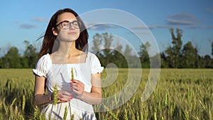 A young woman in a white dress stands in a green wheat field and holds a spikelet in her hands. The girl touches the