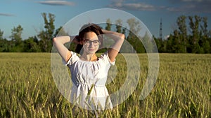 A young woman in a white dress stands in a green wheat field and touches her hair. Girl enjoys the sun in the field.