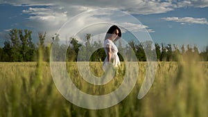 A young woman in a white dress walks on a green wheat field. View of the girl through the spikelets.