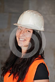 Young woman in white hard hat and orange high visibility jacket, long dark hair, looking into camera and smiling. Closeup detail