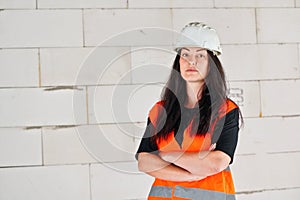 Young woman in white hard hat and orange high visibility vest, long dark hair, hands crossed, looking into camera. Blurred