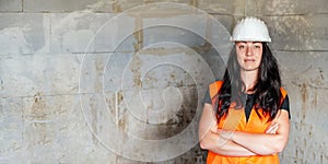 Young woman in white hard hat and orange high visibility vest, long dark hair, hands crossed, looking confident. Blurred