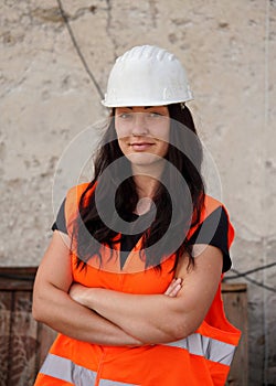 Young woman in white hard hat and orange high visibility vest, long dark hair, hands crossed, looking confident. Blurred