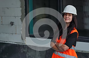 Young woman in white hard hat and orange high visibility vest, long dark hair, looking into camera, hands crossed confident.