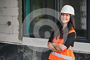 Young woman in white hard hat and orange high visibility vest, long dark hair, looking into camera, hands crossed confident.