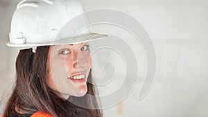 Young woman in white hard hat and orange high visibility vest, long dark hair, looking over her shoulder into camera, smiling.