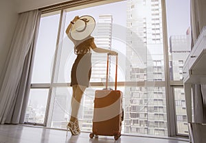 Young  woman with white hat and holding the luggage which standing nearly window, looking to beautiful view of the city