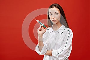 Young woman in white lab coat with a syringe in hands, isolated on red background. Young nurse have prepared a syringe