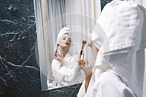 Young woman in white lab coat and towel on her head using a brush in front of a make-up mirror