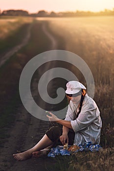 Young woman in white man& x27;s shirt and cap sits in a wheat field near the road on the ground and has lunch