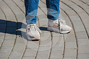 A young woman in white and pink sneakers and blue jeans is standing on the paved sidewalk. Summer walking, entertainment and