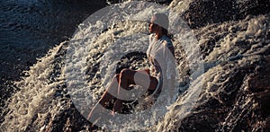 Young woman in white shirt and bikini sits on rock in water flow