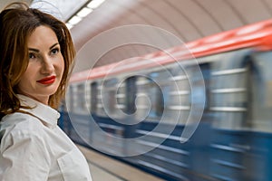 Young woman in white shirt with red hair and red lipstick stands on subway platform against background of passing train