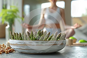 A young woman in a white sports bra and leggings meditates and doing yoga in a sunny room, with a bowl of asparagus and