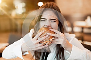 Young woman in white stylish blouse biting with appetite fresh tasty burger during lunch in trendy cafe feeling ultimate hunger