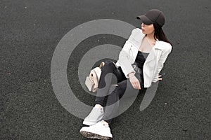 Young woman in white stylish leather jacket in a t-shirt in ripped jeans in white sneakers in a trendy baseball cap is resting