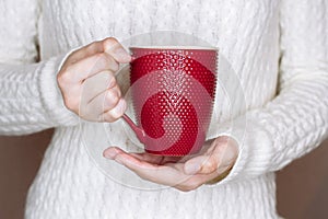 Young woman in white sweater is holding red cup of tea or coffee