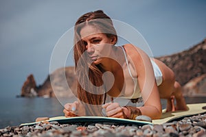 Young woman in white swimsuit with long hair practicing stretching outdoors on yoga mat by the sea on a sunny day. Women