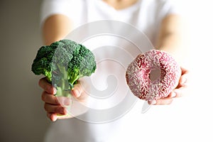 Young woman in white T-shirt choosing between broccoli or junk food, donut. Healthy clean detox eating concept photo
