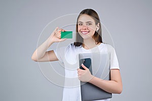 Young woman in white t-shirt holding credit card near face and laptop, isolated over gray background. Pay wit credit or
