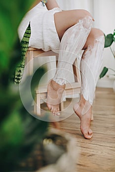 Young woman in white towel applying shaving cream on her legs in home bathroom with green plants. Skin care and wellness. Hair